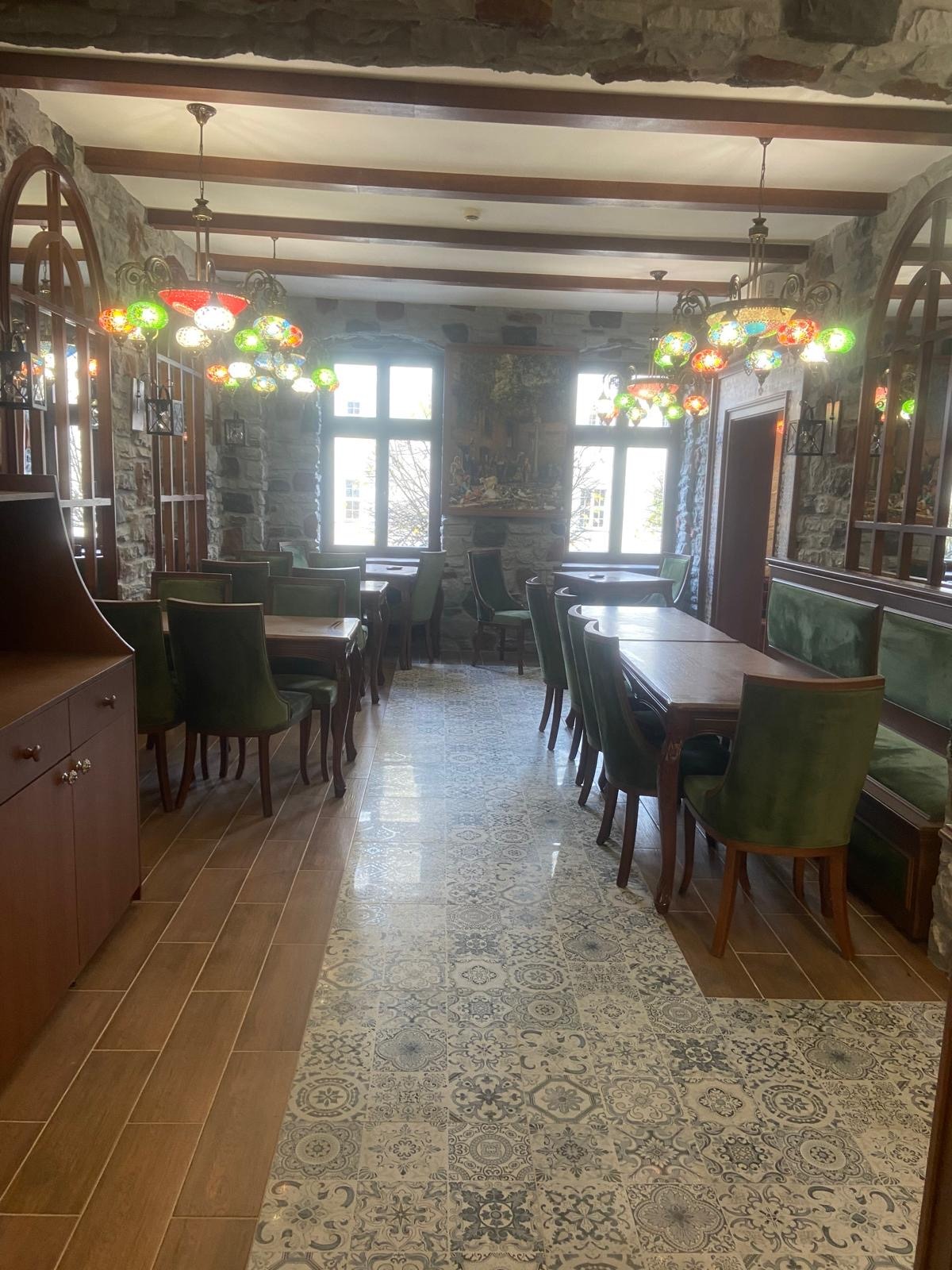 Wnętrze restauracji z kamiennymi ścianami, drewnianymi belkami stropowymi, mozaikową podłogą i kolorowymi lampami wiszącymi.