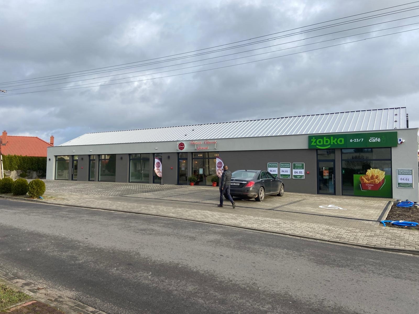 Nowoczesny, parterowy budynek handlowo-usługowy z szarą elewacją i białym dachem, widoczne szyldy 'Żabka' i 'Mikro-Cukiernia Głogówek', zaparkowany samochód osobowy i przechodzący mężczyźni.