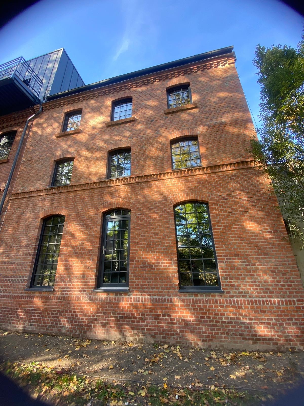 Trzykondygnacyjny budynek z czerwonej cegły z czarnymi, prostokątnymi oknami i drzwiami, widziany pod kątem, z opadłymi liśćmi na ziemi.