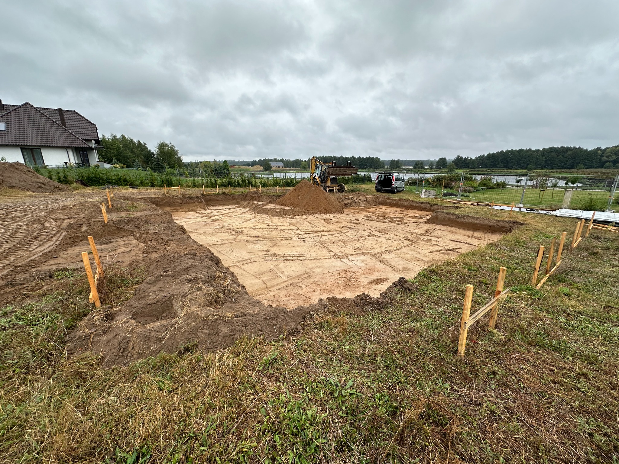 Przygotowany wykop pod fundamenty domu z widocznymi śladami po pracy koparki, drewnianymi słupkami wyznaczającymi obrys i stertą ziemi w tle, pochmurne niebo.