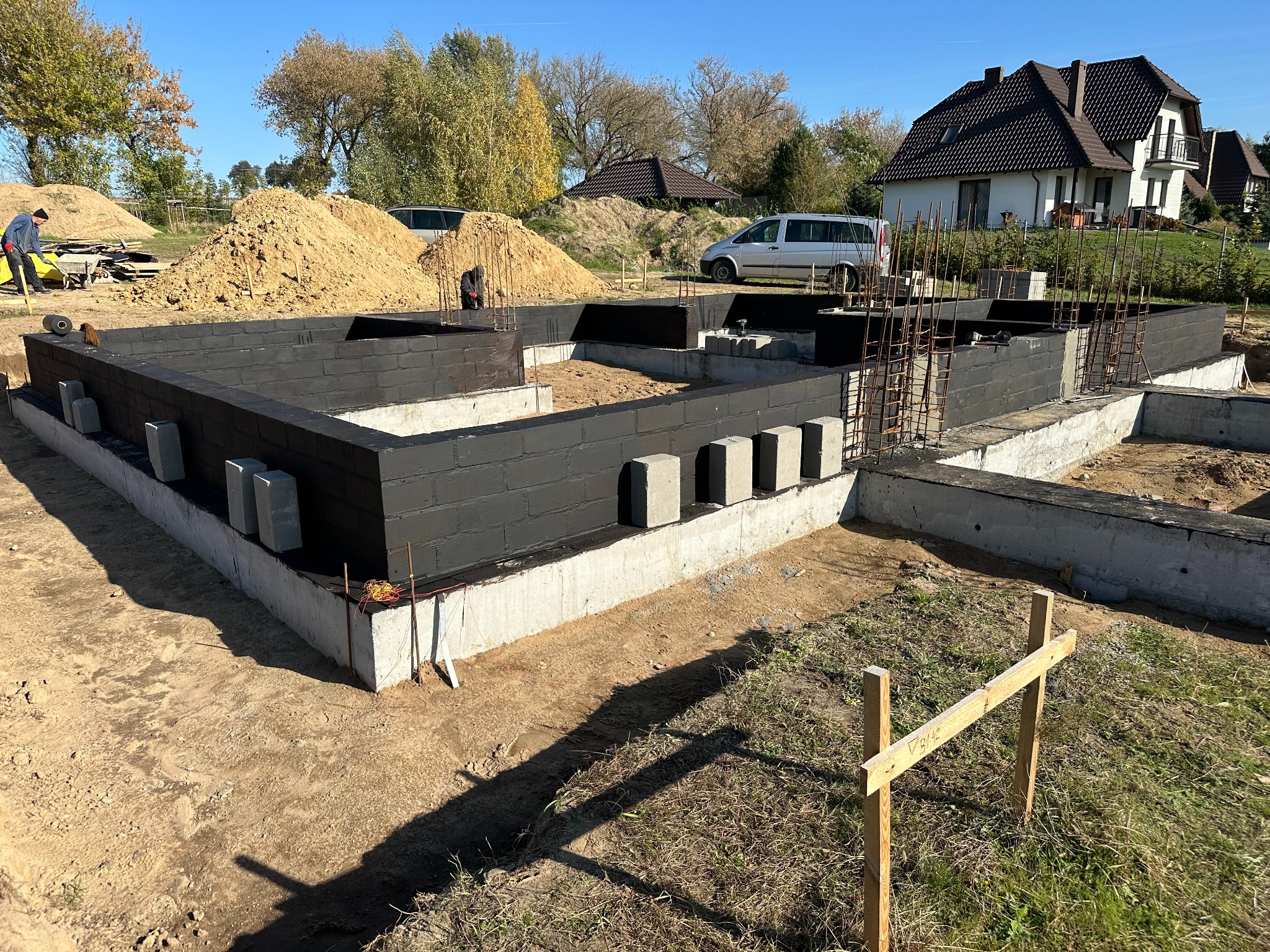Fundamenty domu w trakcie budowy, z czarną izolacją przeciwwilgociową, widoczne zbrojenia i przygotowany teren, w tle kopiec ziemi i budynek mieszkalny.