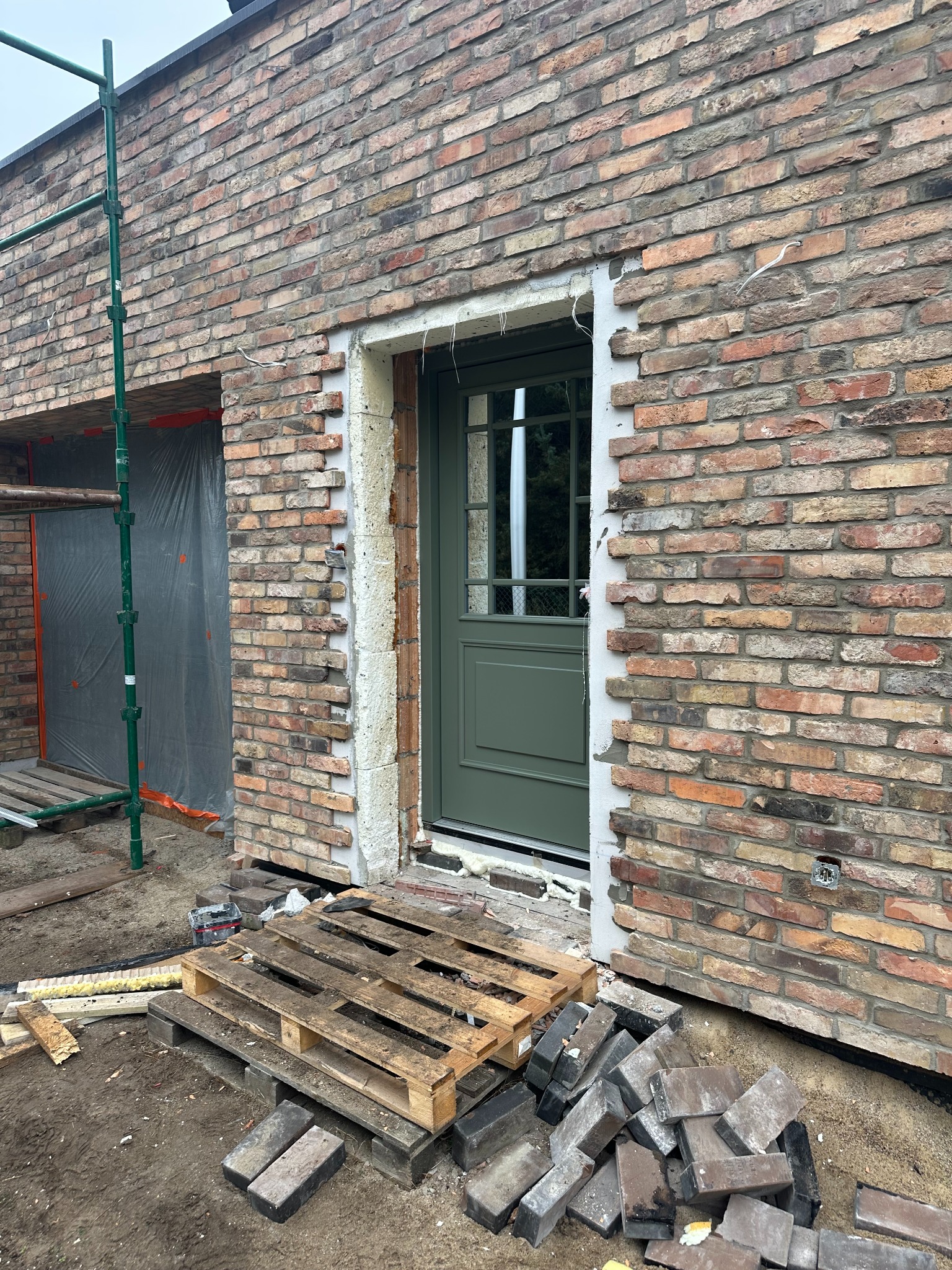 Murowana ściana budynku z zamontowanymi oknami i drzwiami w kolorze oliwkowym, widoczne rusztowanie oraz paleta z materiałami budowlanymi, etap prac wykończeniowych.