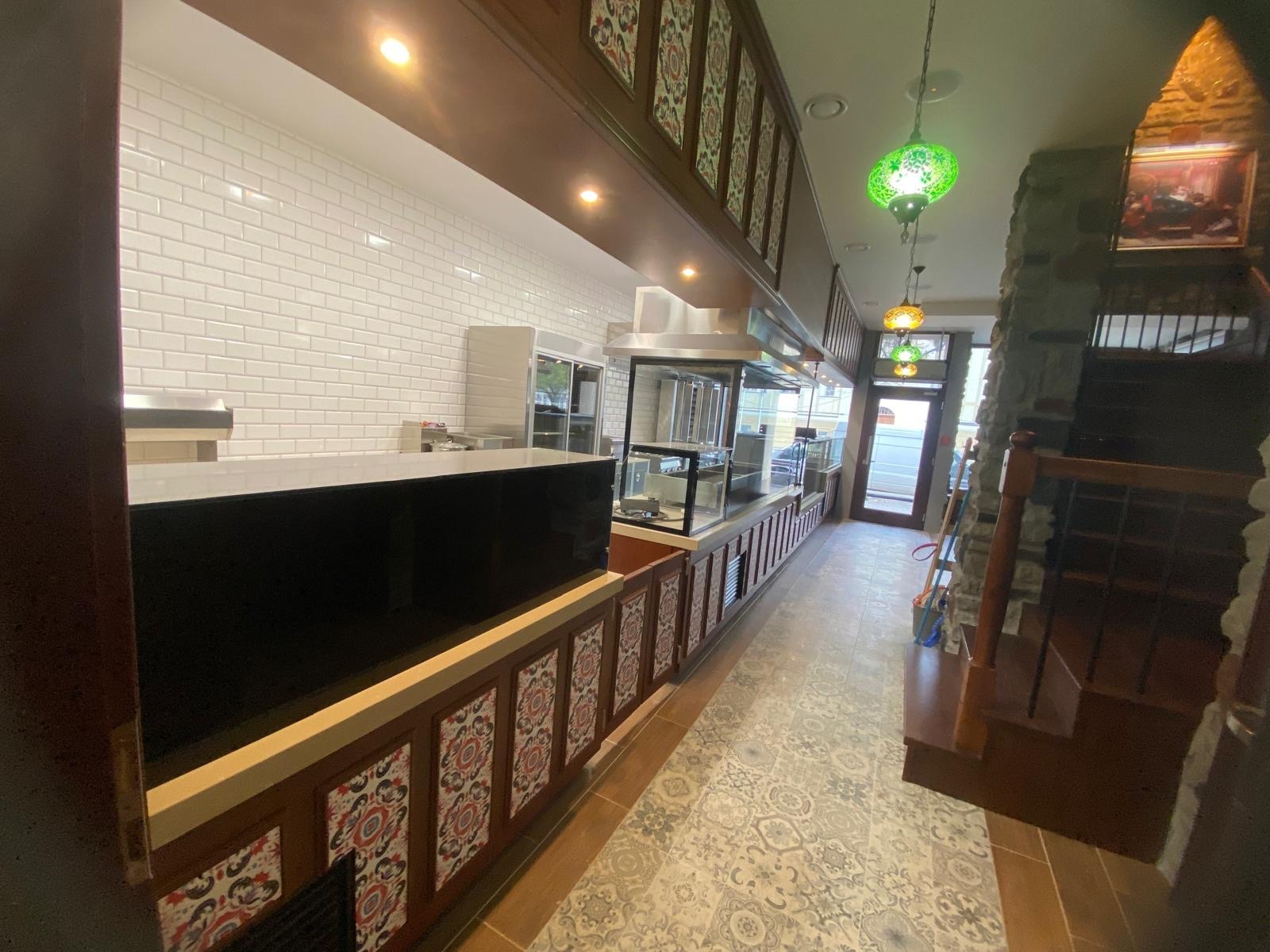 Wnętrze lokalu gastronomicznego z białymi, prostokątnymi płytkami na ścianie, drewnianymi elementami dekoracyjnymi z ornamentami, dekoracyjnymi lampami wiszącymi i kamienną ścianą z schodami.