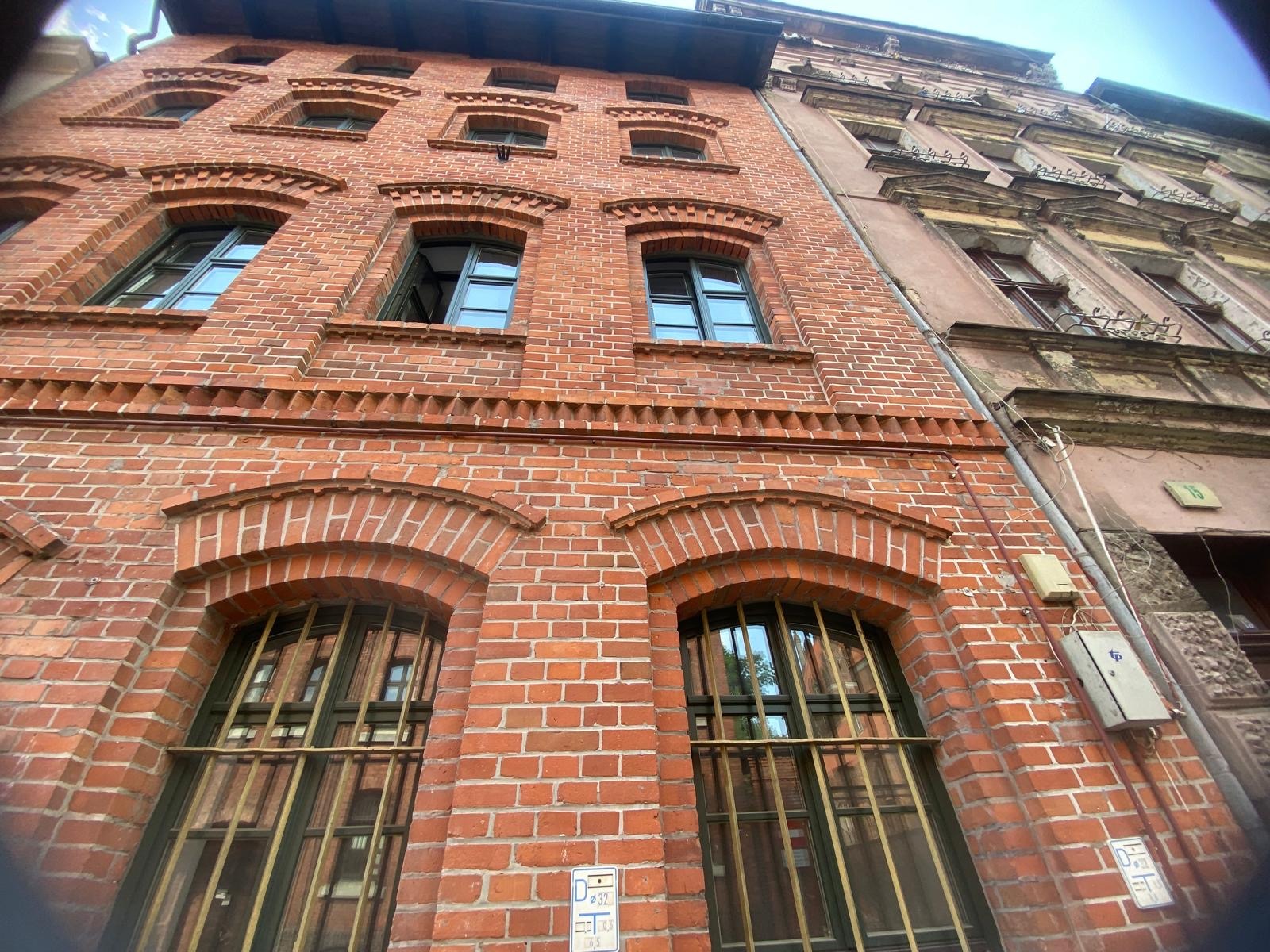 Fasada budynku z czerwonej cegły z oknami zabezpieczonymi kratami, widziana z dołu pod kątem, obok fragmentu elewacji innego budynku.