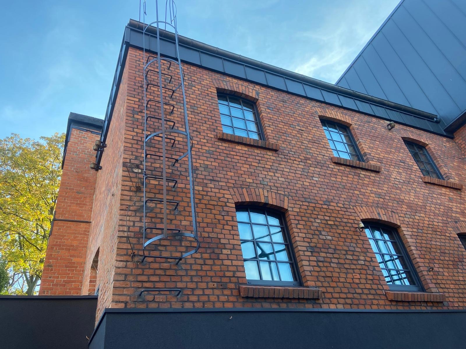Ceglana elewacja budynku z oknami w czarnych ramach i metalową drabiną przymocowaną do ściany, widok z dołu