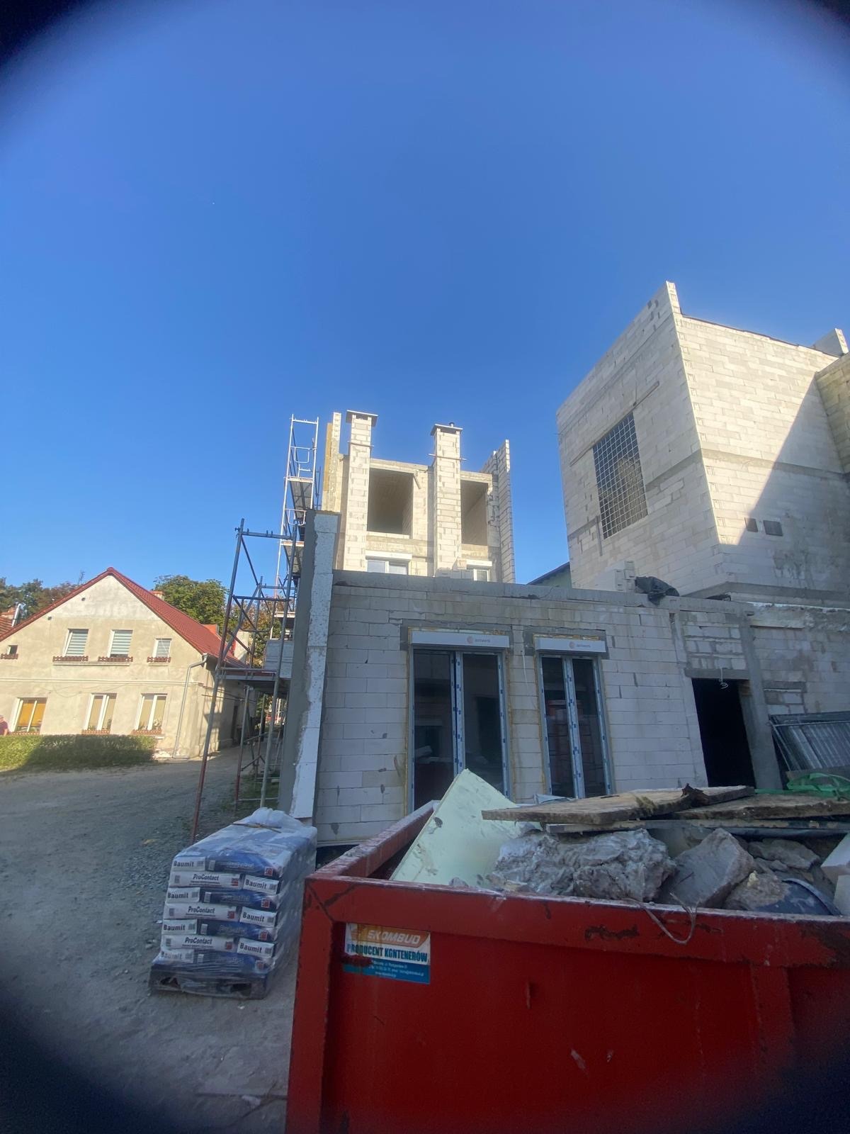 Wznoszenie domu z bloczków, widoczne rusztowania, okna z roletami zewnętrznymi, paleta z materiałami budowlanymi i kontener na gruz na pierwszym planie.