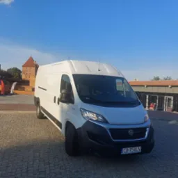 Biały samochód dostawczy Fiat Ducato zaparkowany na brukowanym placu z widokiem na ceglaną wieżę zamkową w tle pod błękitnym niebem.