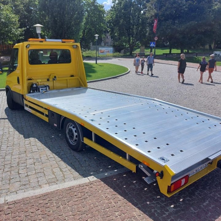 Żółta laweta z aluminiową platformą, zaparkowana na brukowanej drodze, z ludźmi i zielenią w tle.