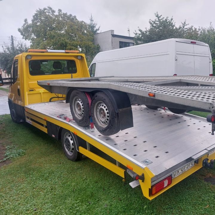 Żółta laweta z platformą do przewozu pojazdów, zaparkowana na trawniku przed białym busem.