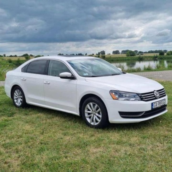 Biały samochód osobowy Volkswagen Passat zaparkowany na trawie z widokiem na rzekę pod pochmurnym niebem.