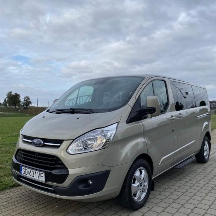 Beżowy Ford Transit Custom Kombi z przyciemnianymi szybami bocznymi i stopniem wejściowym, zaparkowany na kostce brukowej w pochmurny dzień.