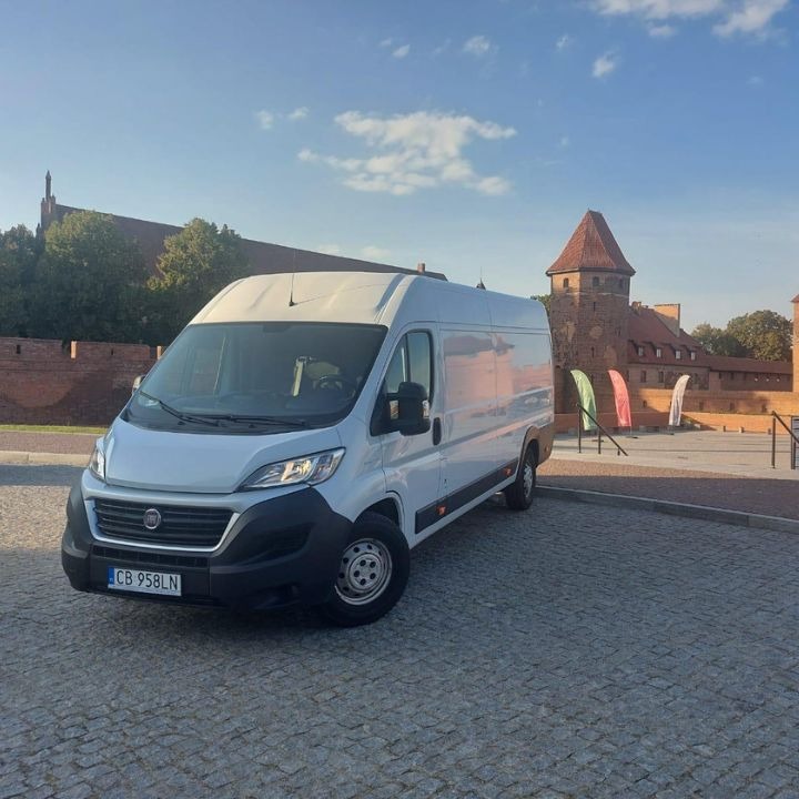 Biały van Fiat Ducato zaparkowany na brukowanym placu z widokiem na Zamek w Malborku w tle, w słoneczny dzień.