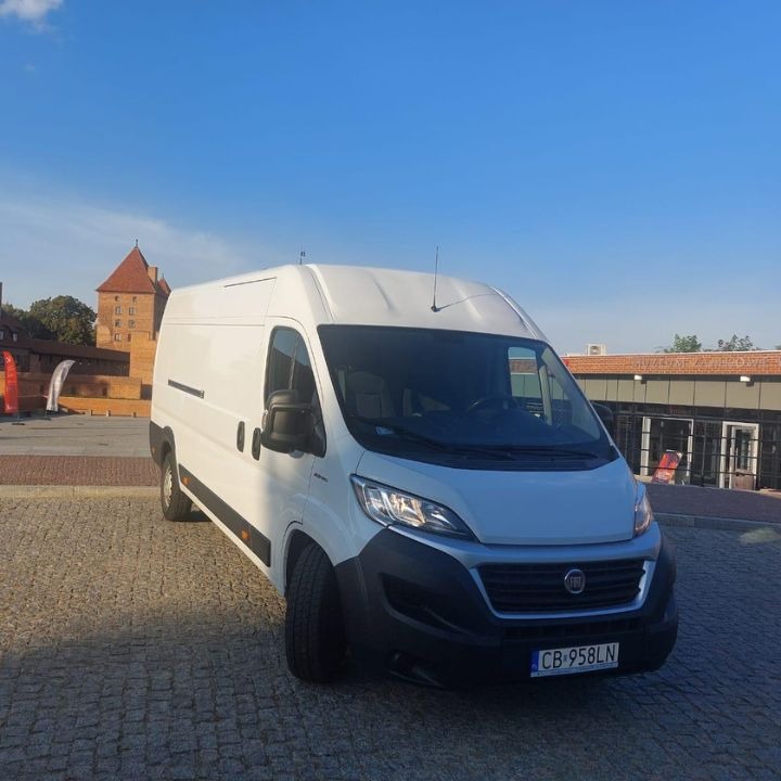 Biały samochód dostawczy Fiat Ducato zaparkowany na brukowanym placu z widokiem na ceglaną wieżę zamkową w tle pod błękitnym niebem.