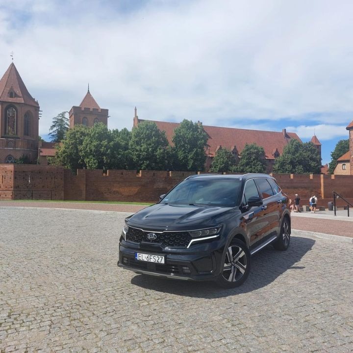 Czarny samochód Kia Sorento zaparkowany na brukowanym placu na tle zamku w Malborku, widoczne tablice rejestracyjne EL 6FS27.