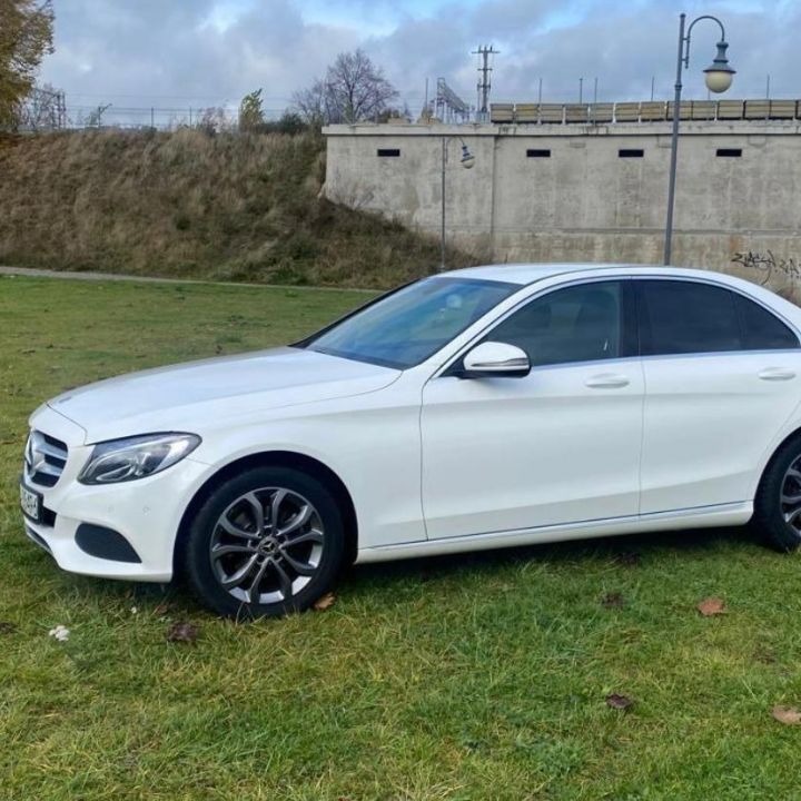 Biały samochód osobowy marki Mercedes-Benz, zaparkowany na trawie przed betonowym murem z graffiti w tle.