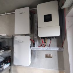 TADEUSZ CHRZĄSZCZ FREE-ENERGY - Zestaw akumulatorów i inwerter fotowoltaiczny CELLX zamontowany na ścianie w pomieszczeniu. Widoczne przewody i elementy instalacji. Ujęcie z dołu.