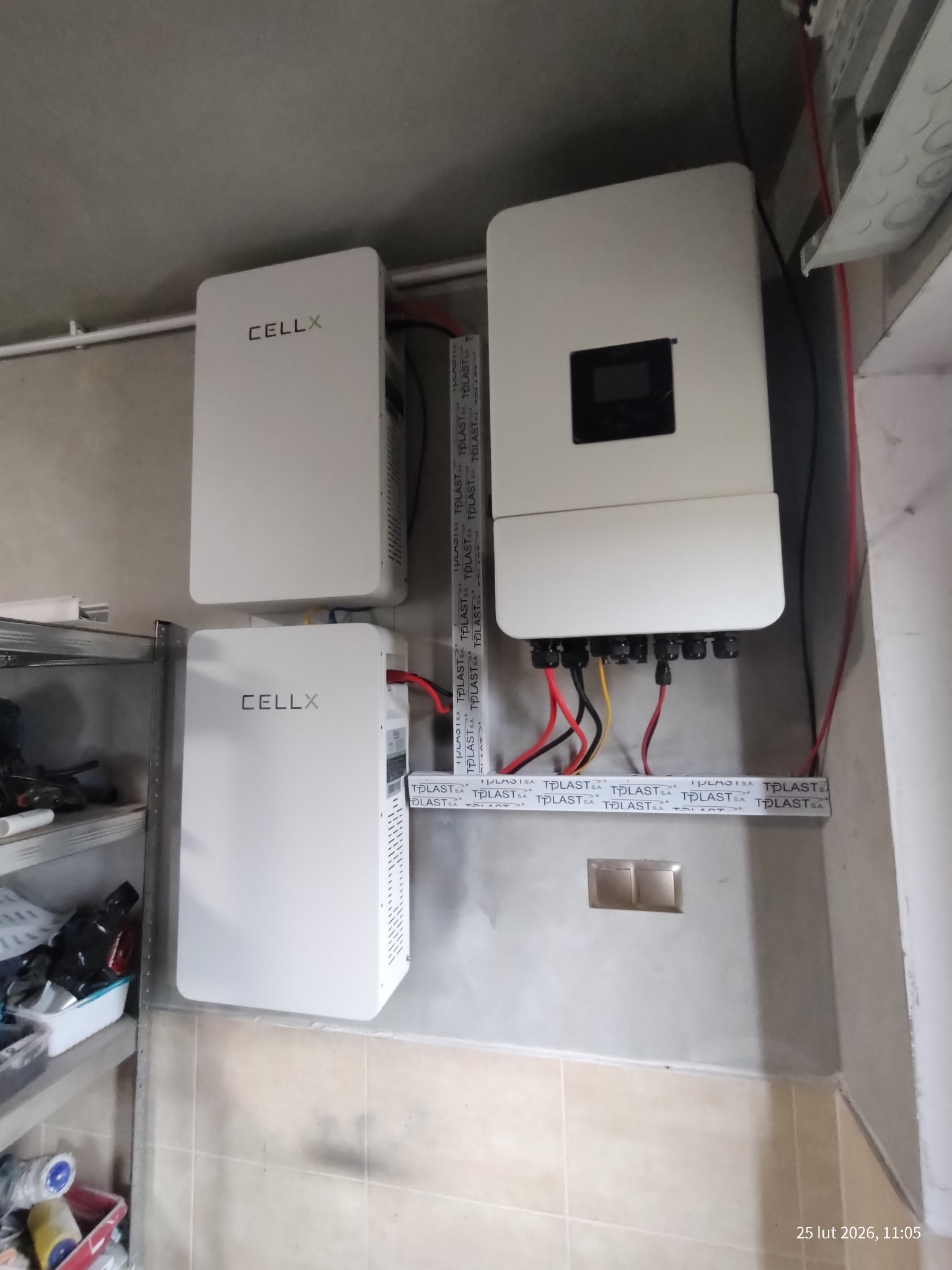 Zestaw akumulatorów i inwerter fotowoltaiczny CELLX zamontowany na ścianie w pomieszczeniu. Widoczne przewody i elementy instalacji. Ujęcie z dołu.