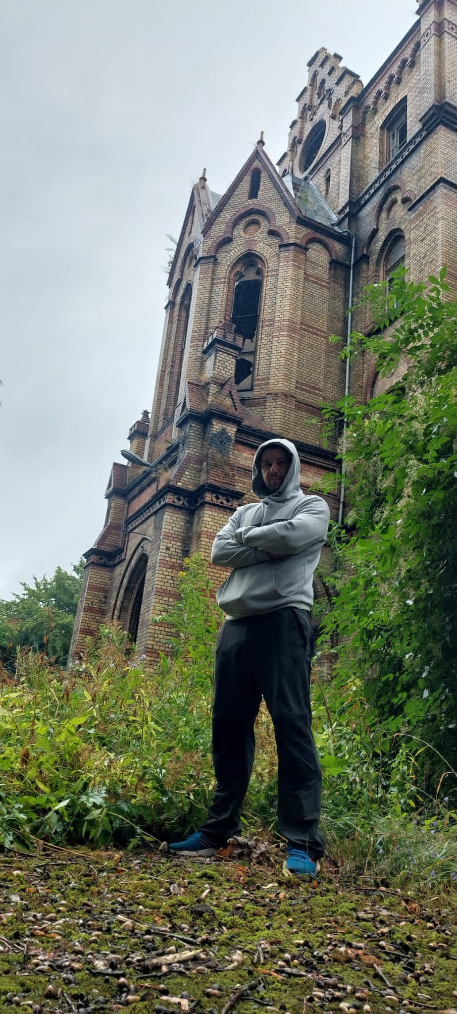 Mężczyzna w bluzie z kapturem stoi na tle zrujnowanego, ceglanego budynku z widocznymi śladami uszkodzeń i braku okien, otoczonego zielenią i chwastami.
