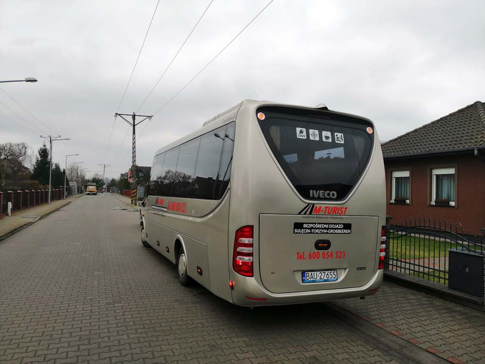 Srebrny autokar marki Iveco z napisem 'M-TURIST' oraz numerem telefonu, zaparkowany na brukowanej ulicy w spokojnej okolicy z domami i słupami elektrycznymi.