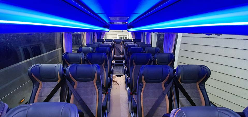 Wnętrze pustego autobusu z rzędami czarnych foteli z brązowymi wstawkami, oświetlone niebieskim światłem LED z góry.