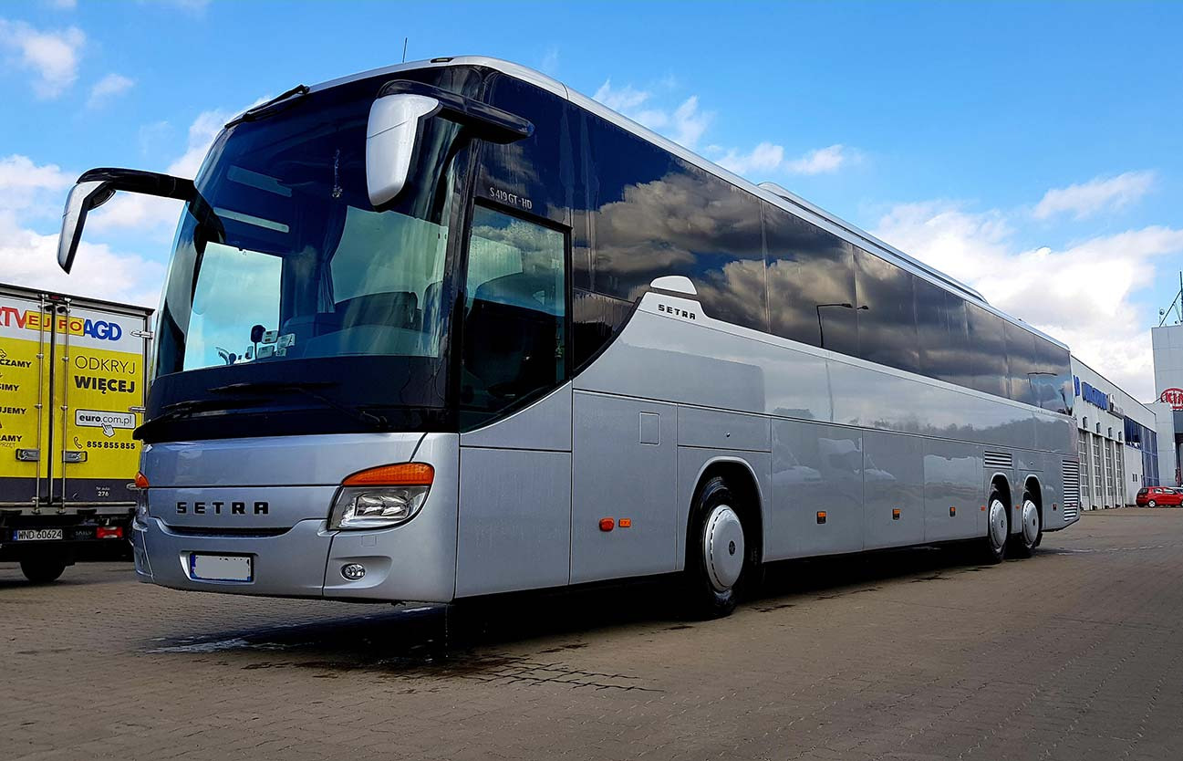 Srebrny autokar Setra S419GT-HD zaparkowany na zewnątrz, z odbiciem chmur w szybach i przyczepą z reklamą w tle.