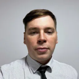 Portret mężczyzny w koszuli w kropki i krawacie na tle jasnej ściany. Ujęcie od przodu.