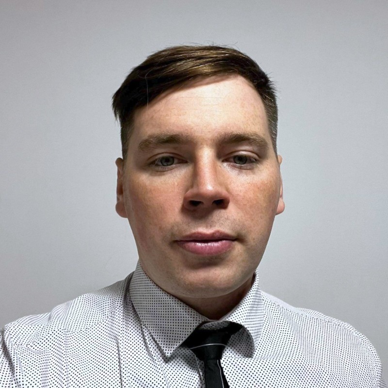 Portret mężczyzny w koszuli w kropki i krawacie na tle jasnej ściany. Ujęcie od przodu.