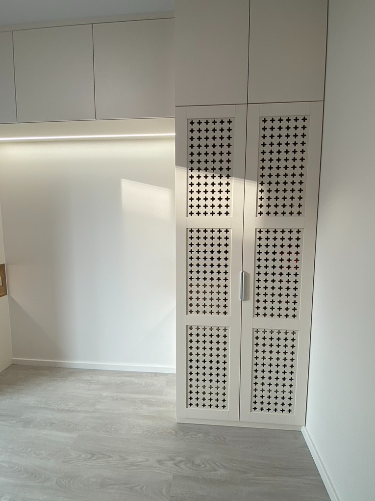 Biała, wysoka zabudowa meblowa w Warszawie z ażurowymi drzwiami szafy, wzór krzyżyków, oświetlenie LED podszafkowe, szare panele podłogowe.