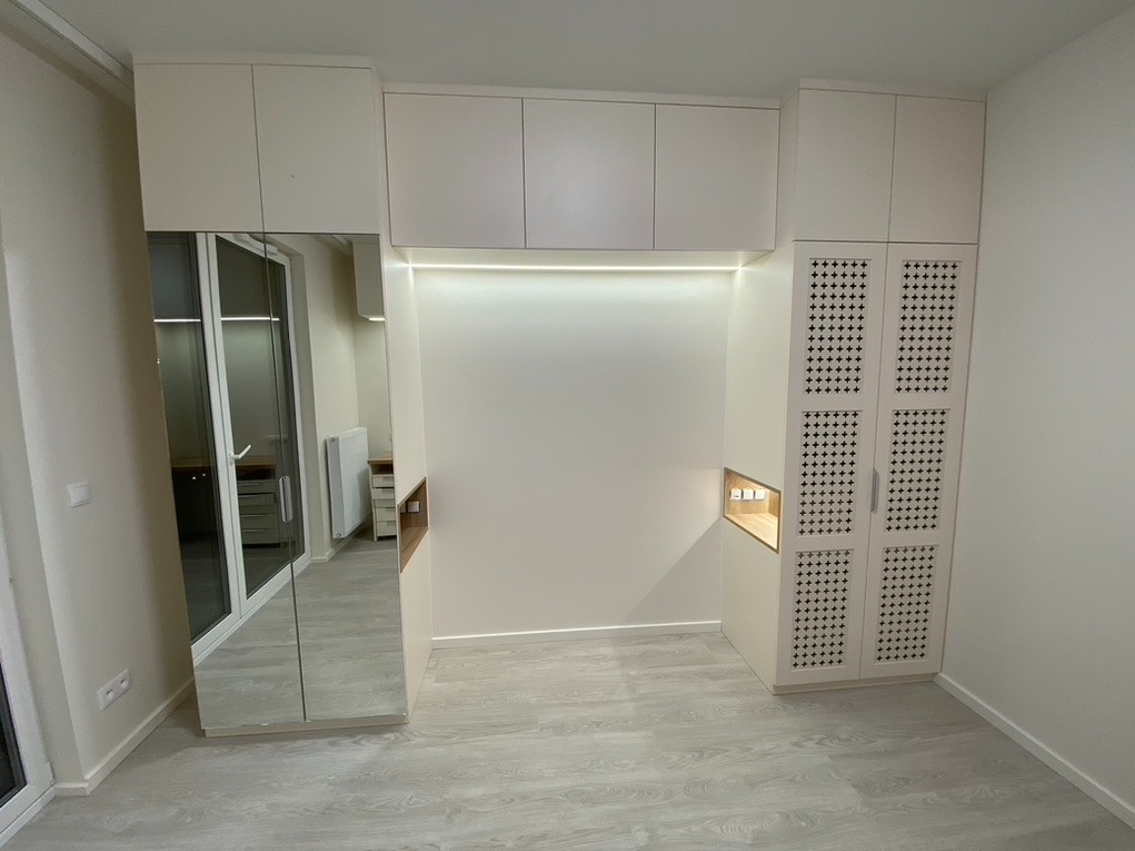 Biała zabudowa meblowa z szafą z lustrzanymi drzwiami i szafkami z ażurowymi frontami, oświetlenie LED wnęki, widok na wnętrze garderoby w lustrze.