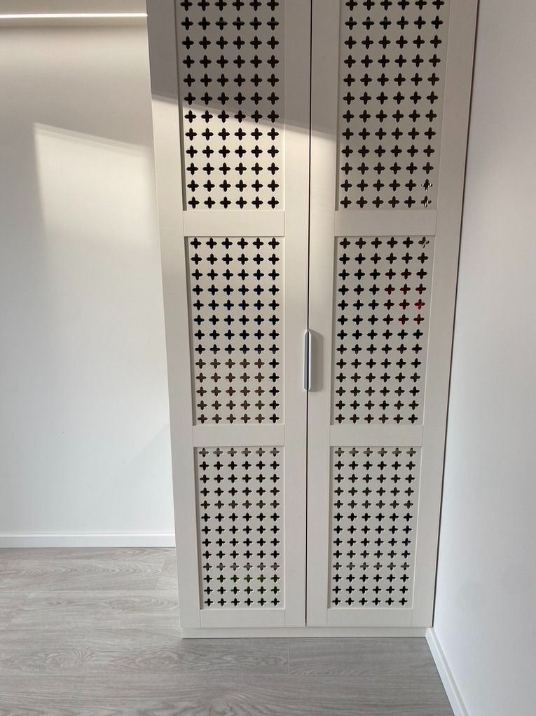 Biała szafa dwudrzwiowa z dekoracyjnymi, ażurowymi frontami w kształcie krzyżyków, na tle białej ściany i szarej podłogi.