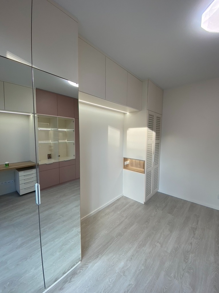 Nowoczesna zabudowa garderoby z białymi szafkami pod sufitem, podświetleniem LED i lustrzanym frontem szafy przesuwnej odbijającym biurko z szufladami w pokoju o jasnej podłodze.