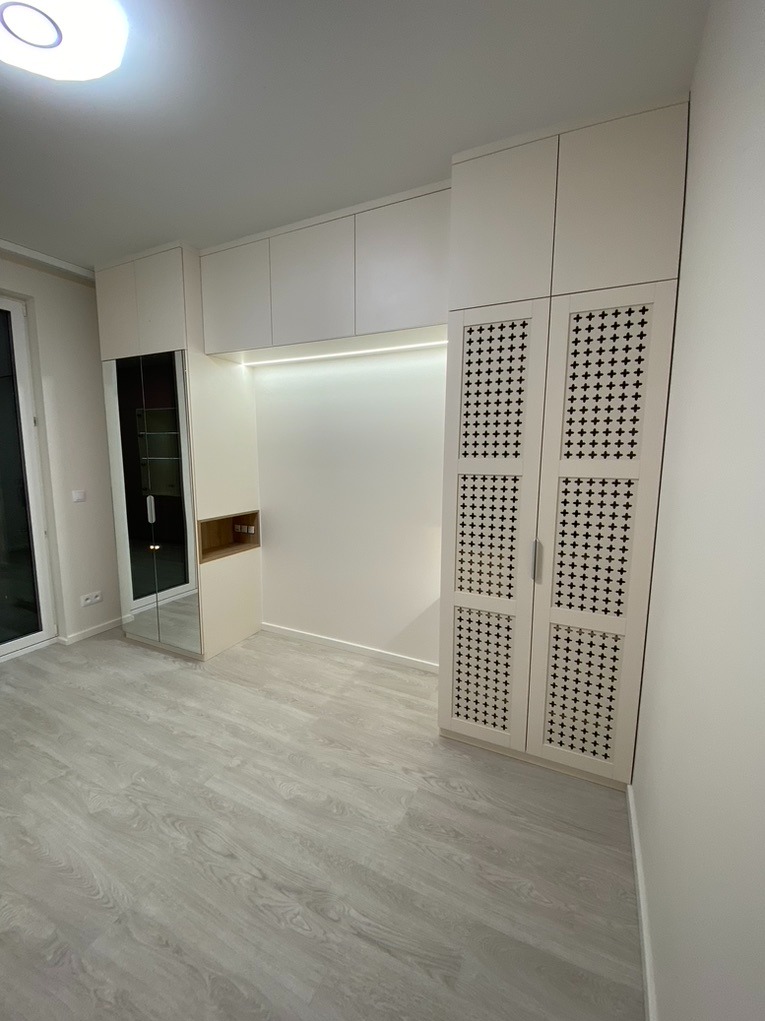 Jasna zabudowa meblowa z szafą z dekoracyjnymi ażurowymi drzwiami w kształcie krzyżyków, nad nią rząd szafek podwieszanych i oświetlenie LED, obok szafa z lustrem, na podłodze panele w odcieniach...