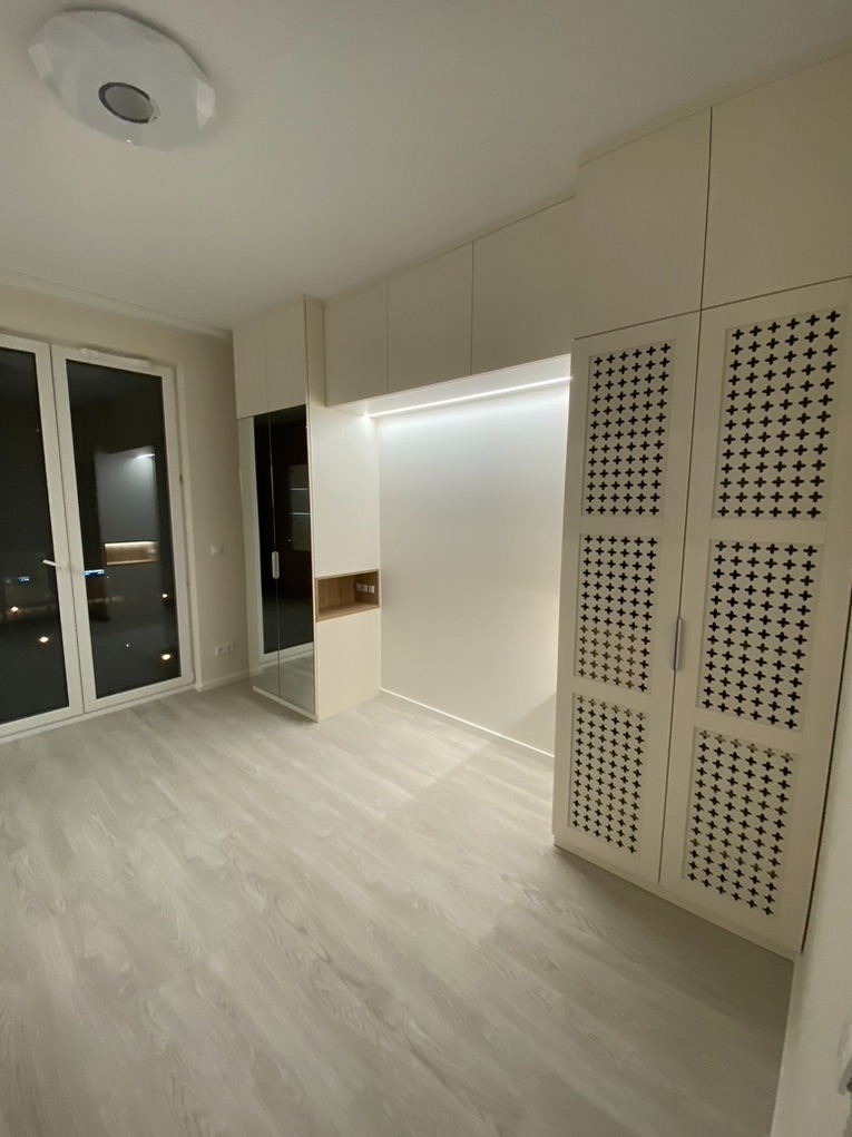 Nowoczesna zabudowa meblowa w jasnym pokoju z dużym oknem balkonowym, obejmująca szafę z dekoracyjnymi ażurowymi drzwiami w kształcie krzyżyków i wbudowane oświetlenie LED.