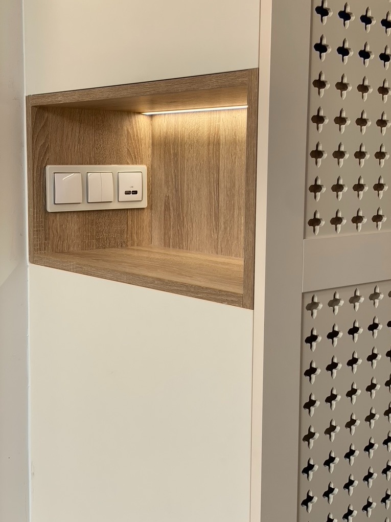 Wnęka oświetlona LED z drewnopodobnym wykończeniem, zawierająca białe włączniki światła i gniazdo USB, sąsiadująca z ażurowym panelem dekoracyjnym.