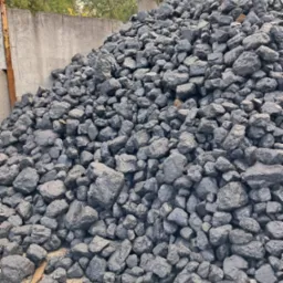 Stos kostki opału o nieregularnych kształtach, ułożony pod betonowym murem z metalową konstrukcją w tle, na tle drzew.