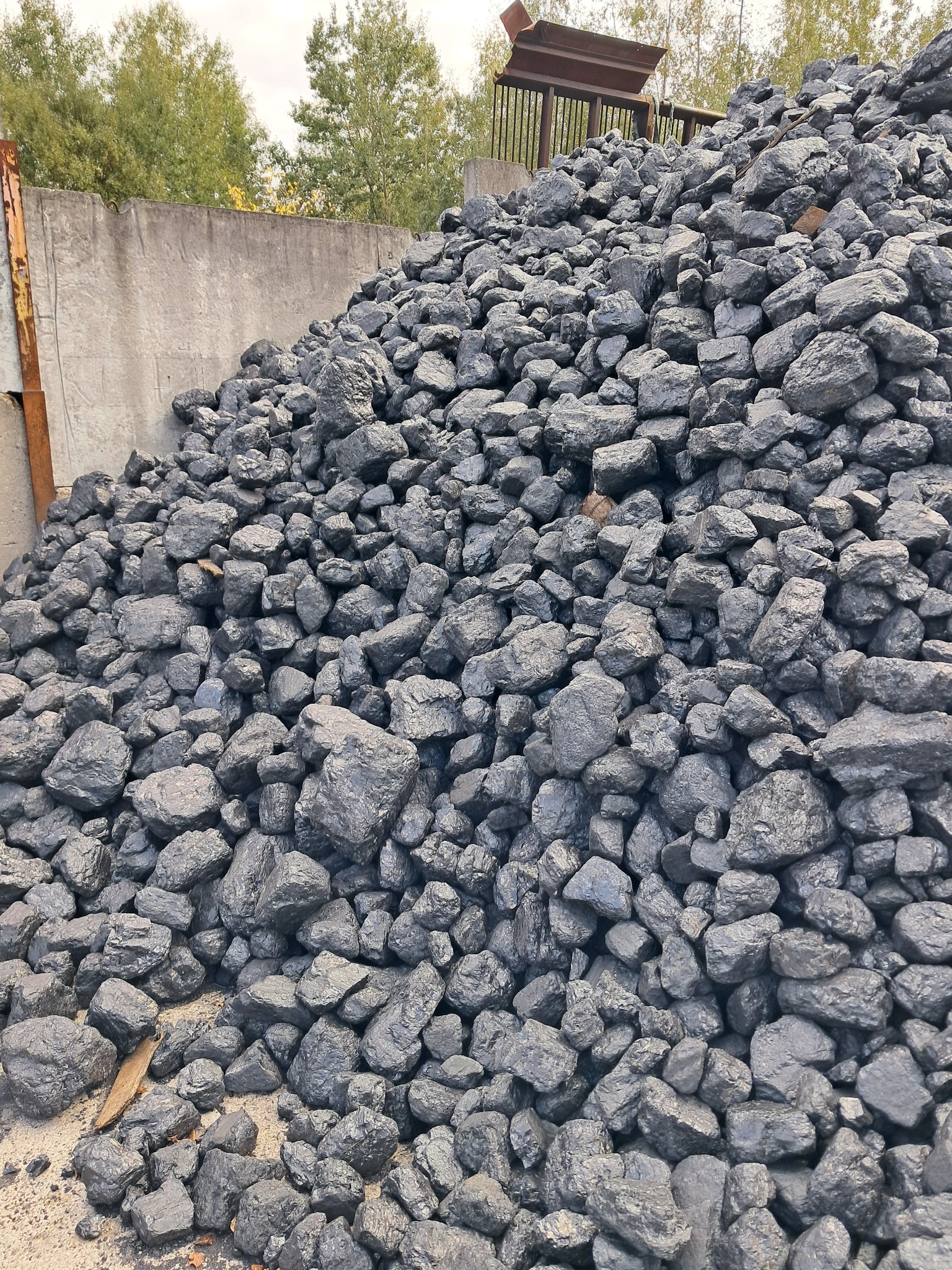 Stos kostki opału o nieregularnych kształtach, ułożony pod betonowym murem z metalową konstrukcją w tle, na tle drzew.