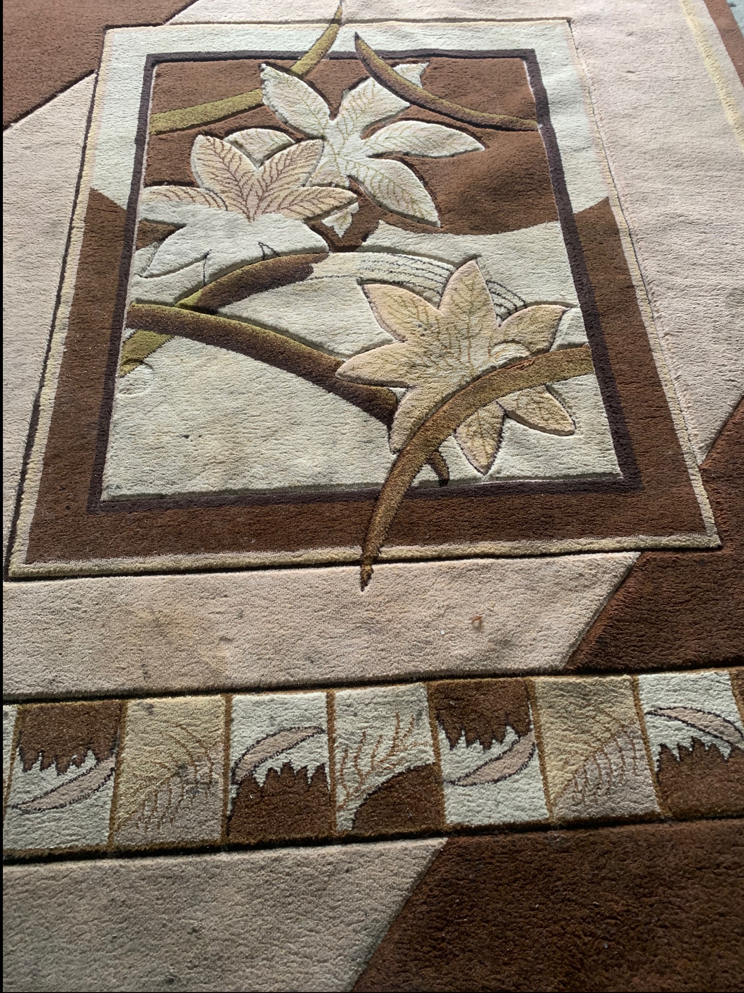 Beżowo-brązowy dywan z motywem liści, widoczne detale wzoru i faktury materiału, delikatne cieniowanie.