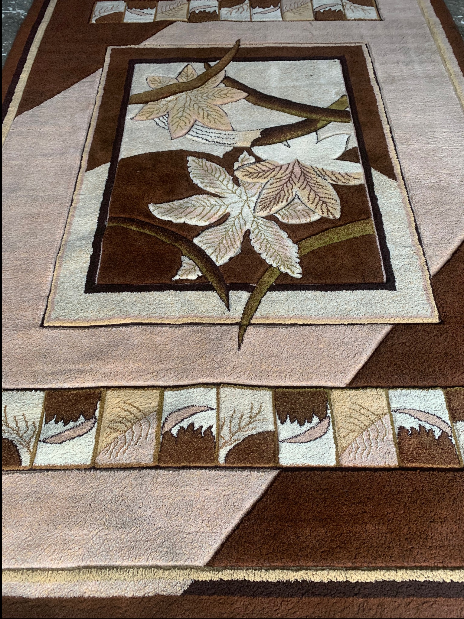 Wzorzysty dywan z motywem kwiatowym w odcieniach brązu i beżu, z centralnym prostokątnym deseniem i geometrycznymi elementami na obrzeżach.