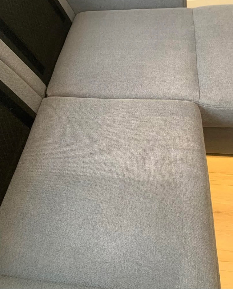 Jasnoszary narożnik z widocznymi śladami po praniu tapicerki, ukazujący różnicę w czystości materiału. Fragment drewnianej podłogi w prawym dolnym rogu.