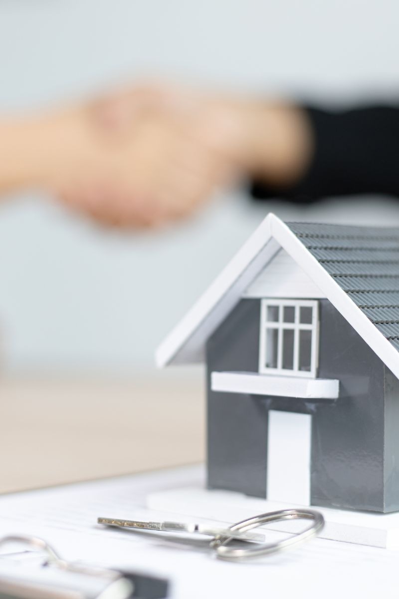 Model domu z kluczami na umowie, w tle uścisk dłoni.