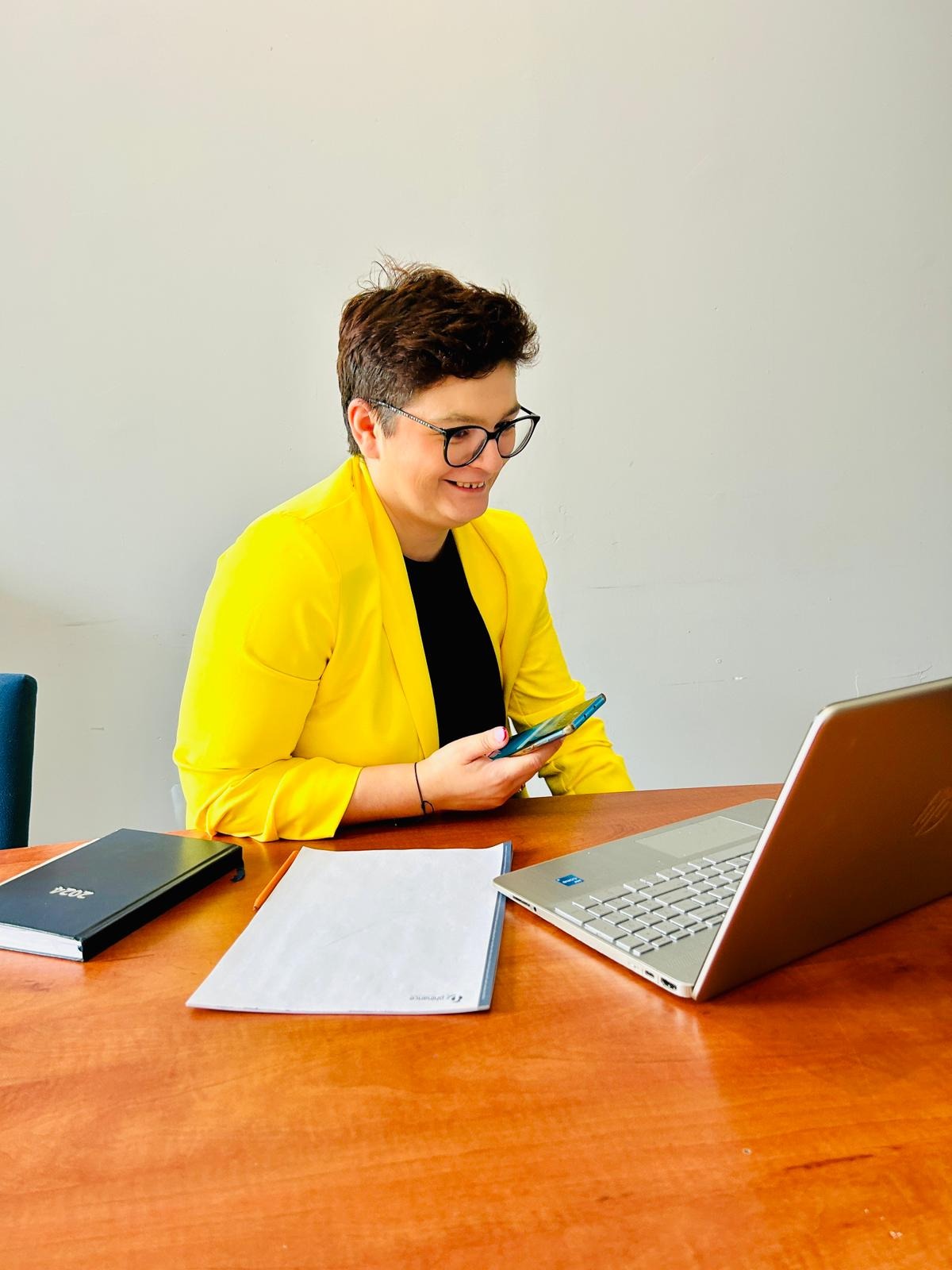 Kobieta w żółtej marynarce, okularach i z krótkimi włosami siedzi przy drewnianym biurku, uśmiecha się i patrzy na smartfon w dłoni, obok leży laptop, notatnik i dokumenty.