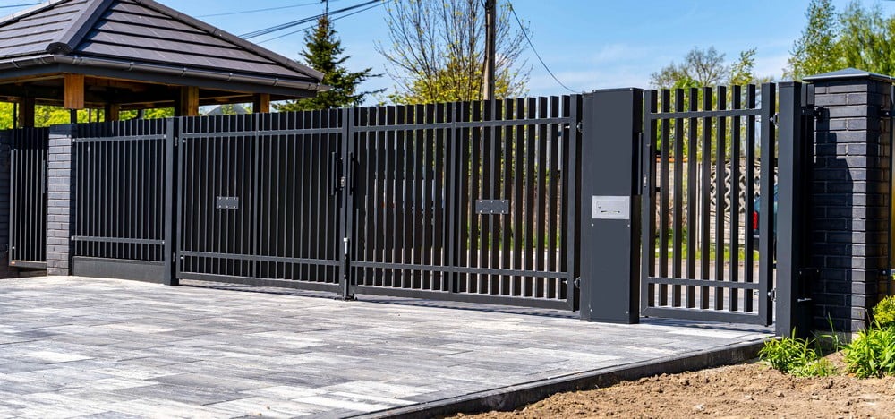 Nowoczesna, metalowa brama wjazdowa z furtką, pomalowana na grafitowo, z pionowymi prętami, zamontowana na posesji z kostką brukową i widoczną altaną w tle.