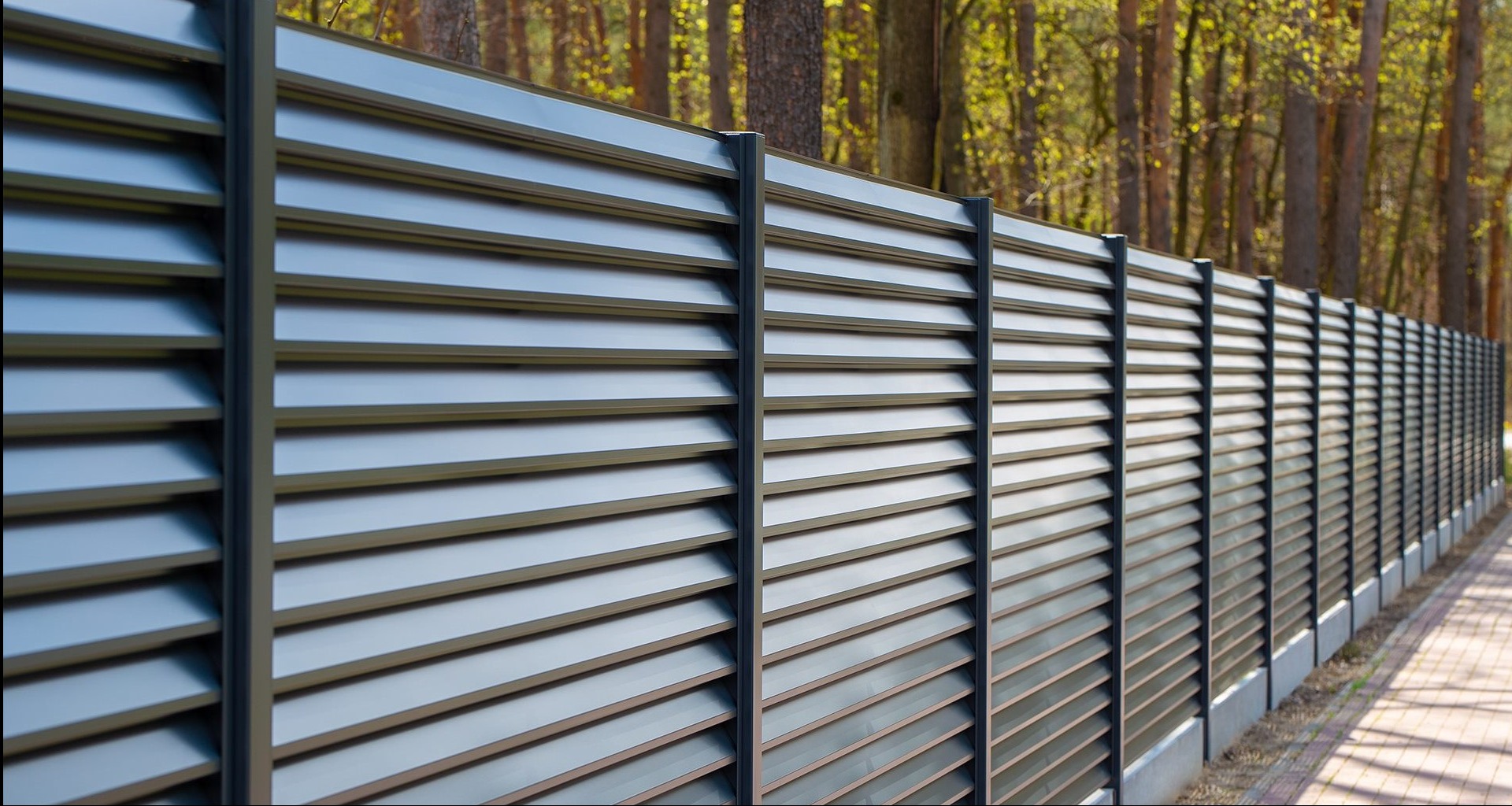 Nowoczesne, szare ogrodzenie metalowe z poziomymi lamelami, zapewniające prywatność, z widocznym fragmentem chodnika i drzewami w tle.
