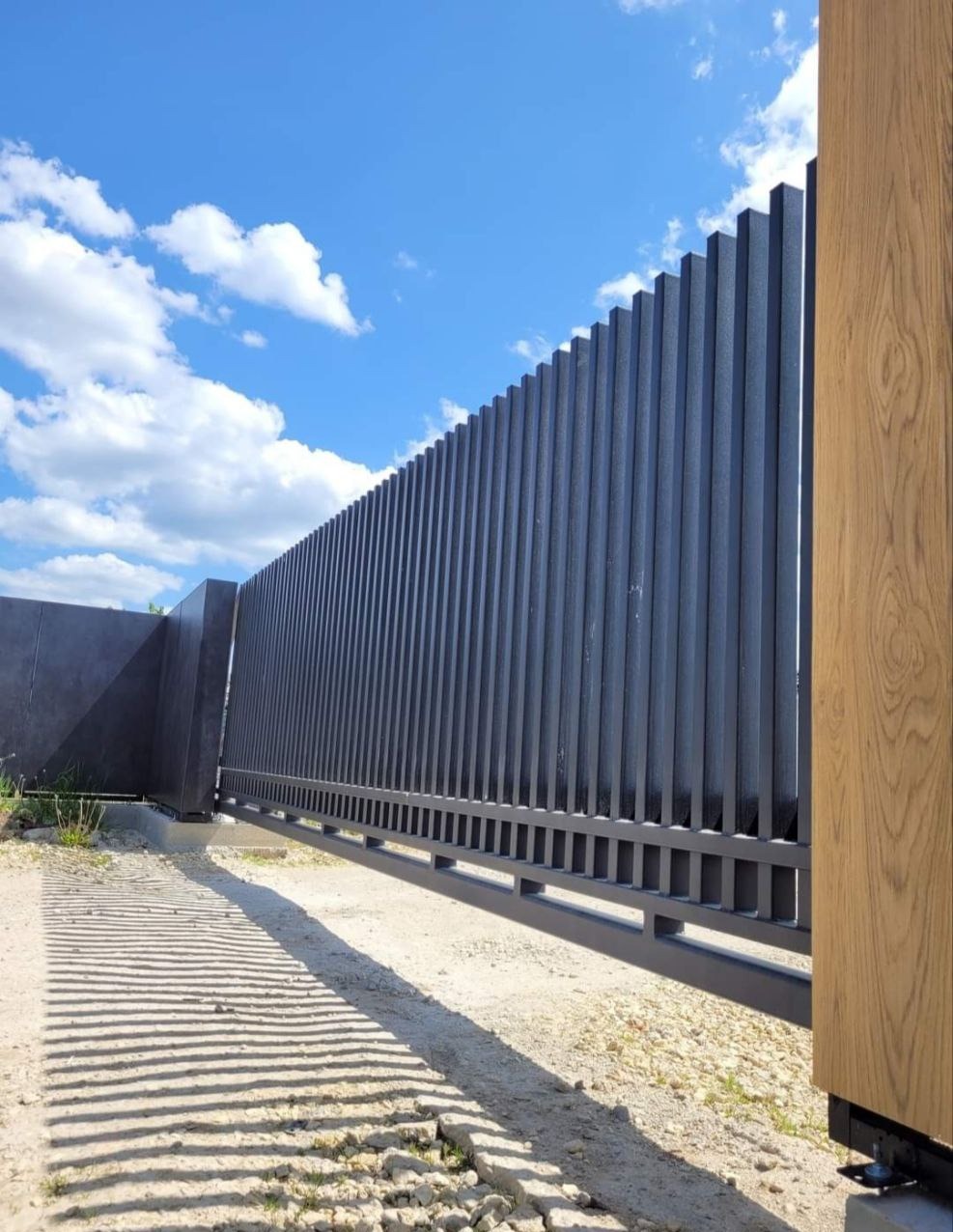 Nowoczesna, przesuwna brama wjazdowa z pionowych, grafitowych profili, widoczna z boku, z elementami imitującymi drewno po prawej stronie. Na ziemi cień rzucany przez bramę.