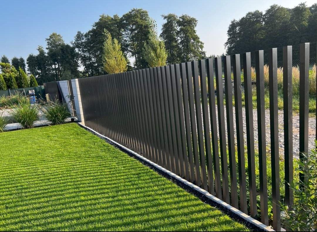 Nowoczesne ogrodzenie metalowe z pionowych, prostokątnych profili w kolorze grafitowym, oddzielające trawnik od ścieżki, z widocznymi drzewami w tle.