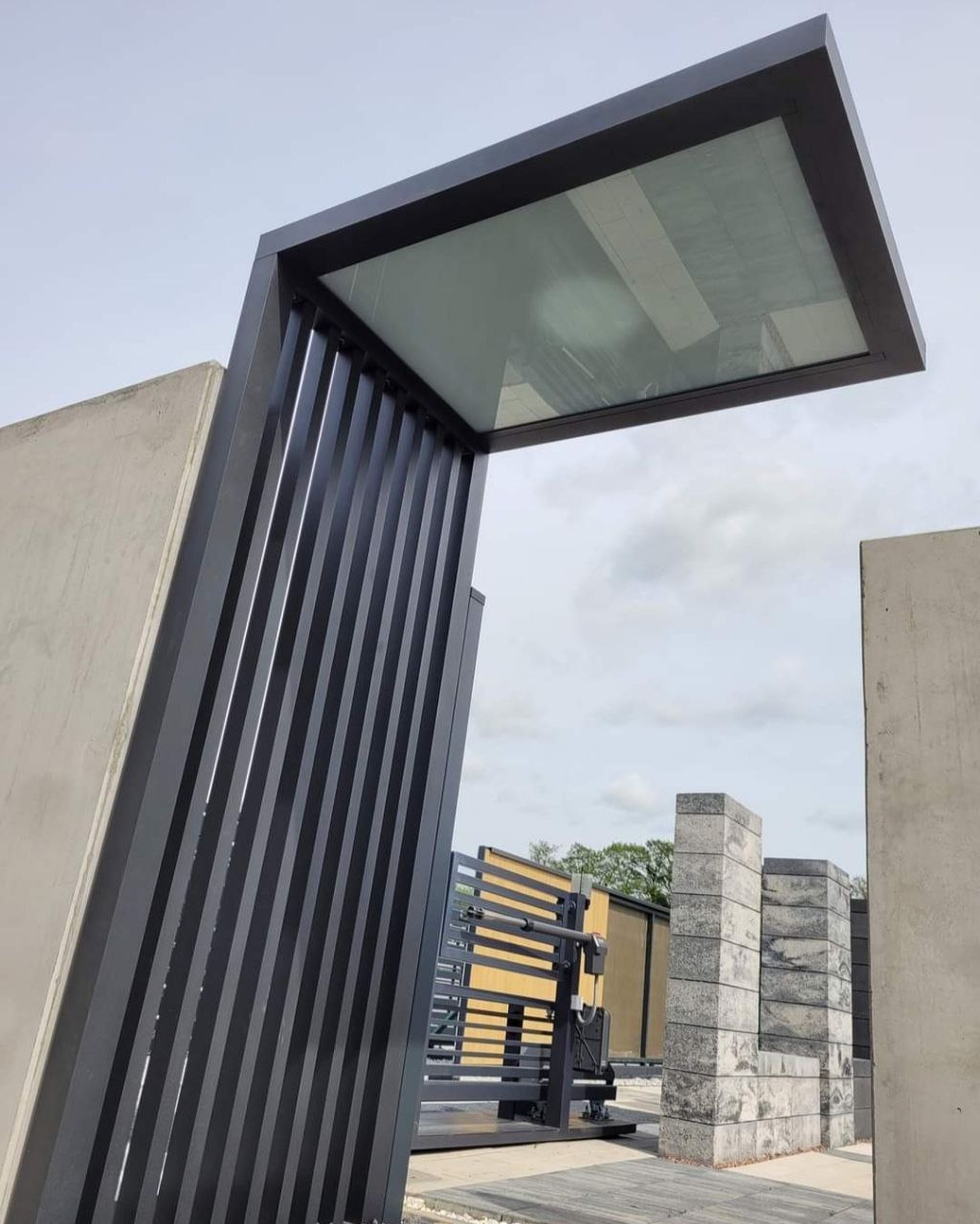 Nowoczesna brama wjazdowa z metalowymi, pionowymi lamelami i zadaszeniem, widok z dołu na tle betonowych słupków i nieba.