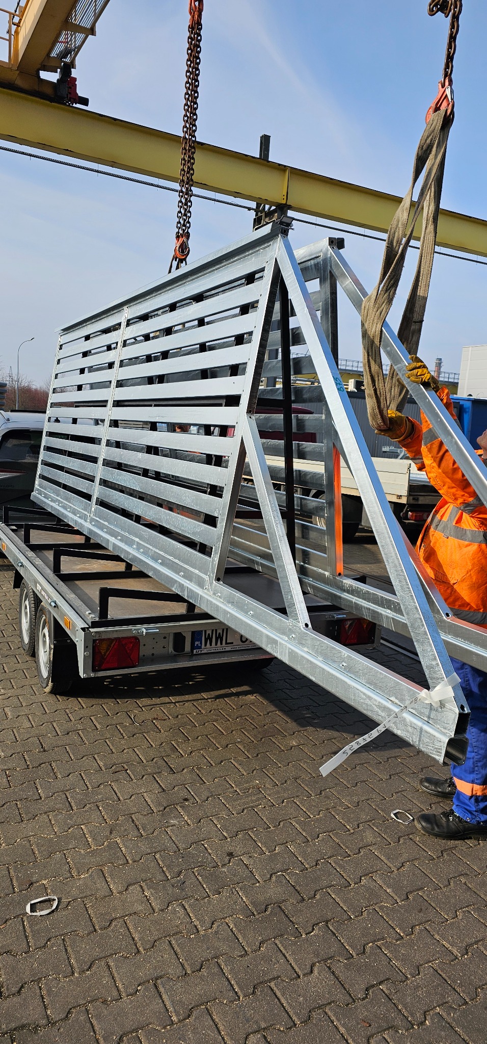 Metalowa konstrukcja bramy wjazdowej z poziomymi szczeblami, unoszona dźwigiem nad przyczepą, pracownik w pomarańczowej kamizelce asekuruje ładunek.