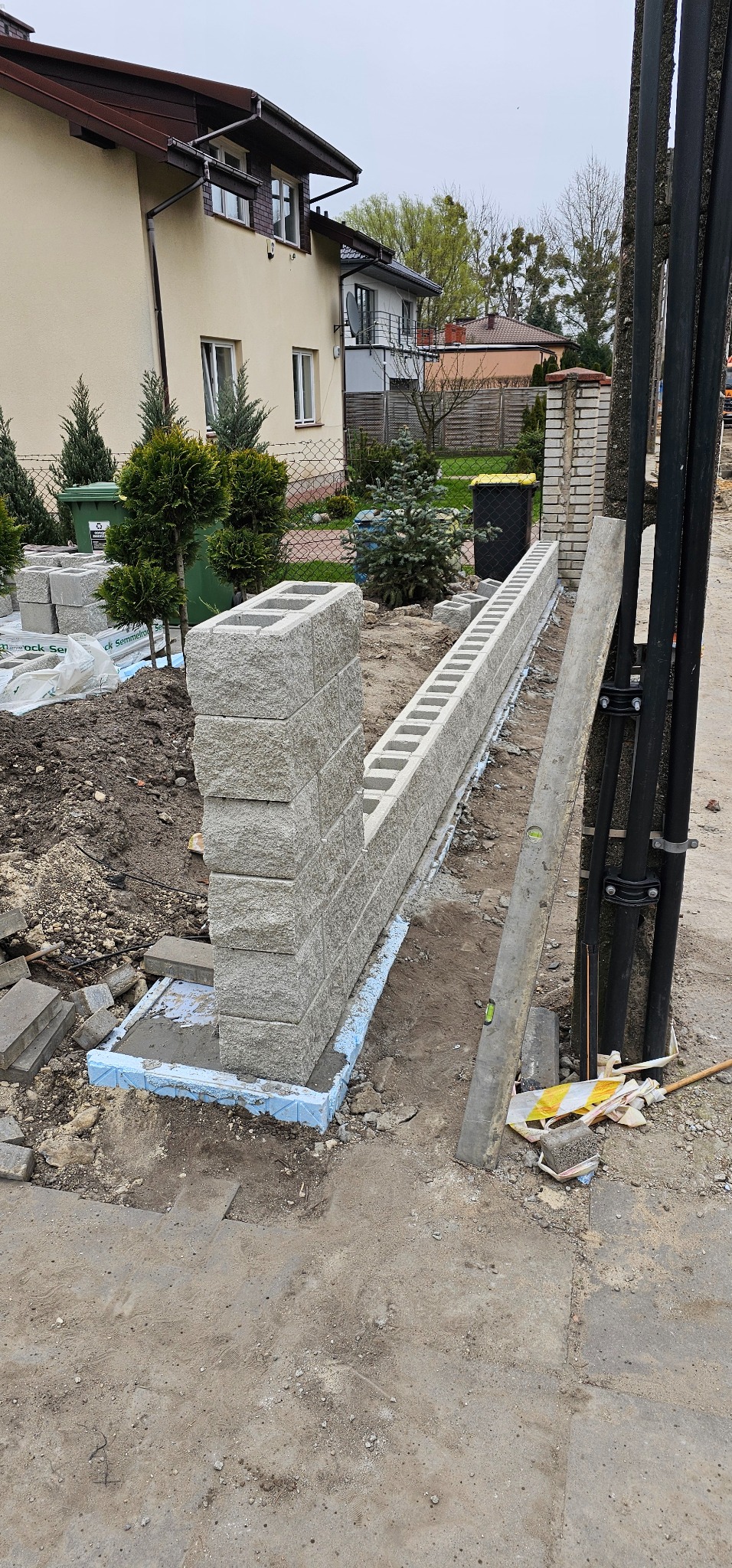Budowa ogrodzenia z szarego bloczka betonowego na fundamencie z izolacją, widoczne narzędzia murarskie i poziomica, w tle domy jednorodzinne.