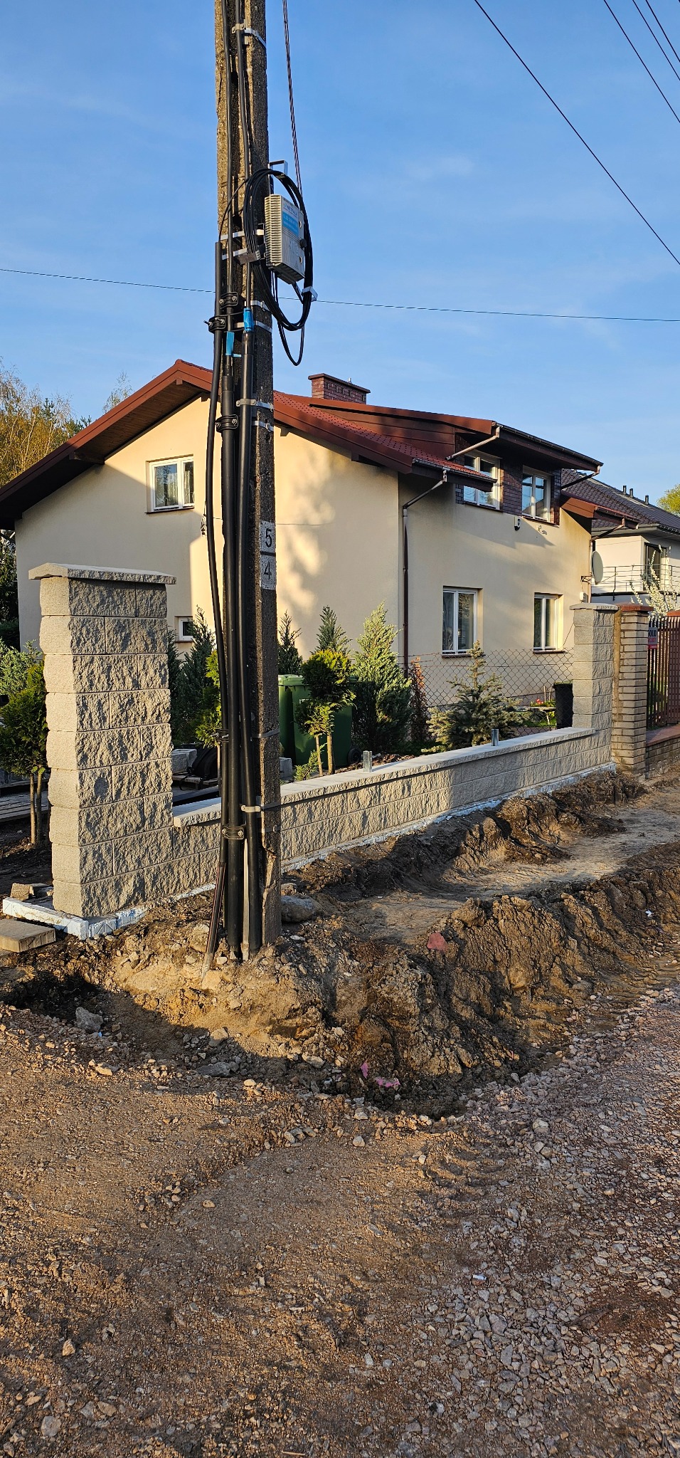 Budowa ogrodzenia z szarej kostki łupanej przed domem jednorodzinnym, widoczne słupki i podmurówka, teren wokół rozkopany, słup energetyczny na pierwszym planie.