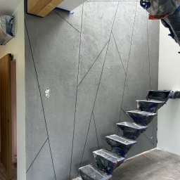 RemontyRs - Ściana w szarym odcieniu z geometrycznym wzorem, zabezpieczone schody i podłoga folią ochronną, widoczny fragment drewnianej belki stropowej i przygotowane gniazdko elektryczne.